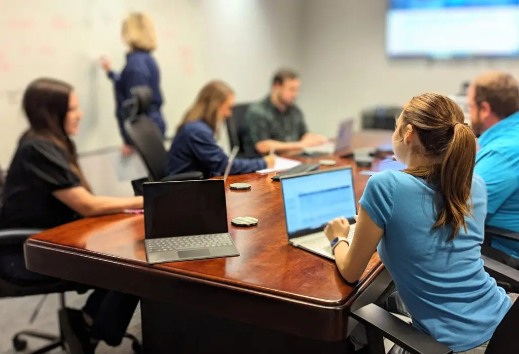 Team meeting in conference room with employees collaborating on laptops and whiteboard.