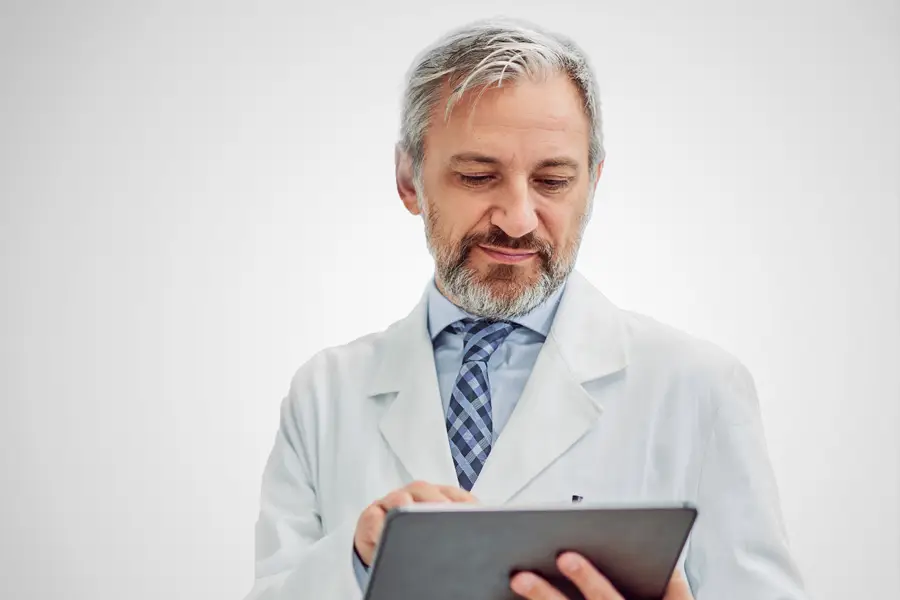 Senior male pharmacist in white lab coat using a digital tablet to review medical or prescription information.