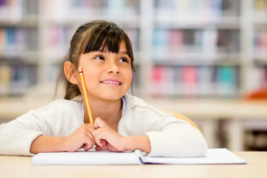 Smiling young girl holding pencil while thinking and writing in notebook at school library