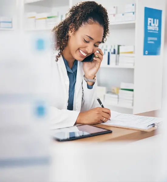 Smiling female pharmacist on phone call while writing notes on a clipboard in a pharmacy with flu shot sign in the background.