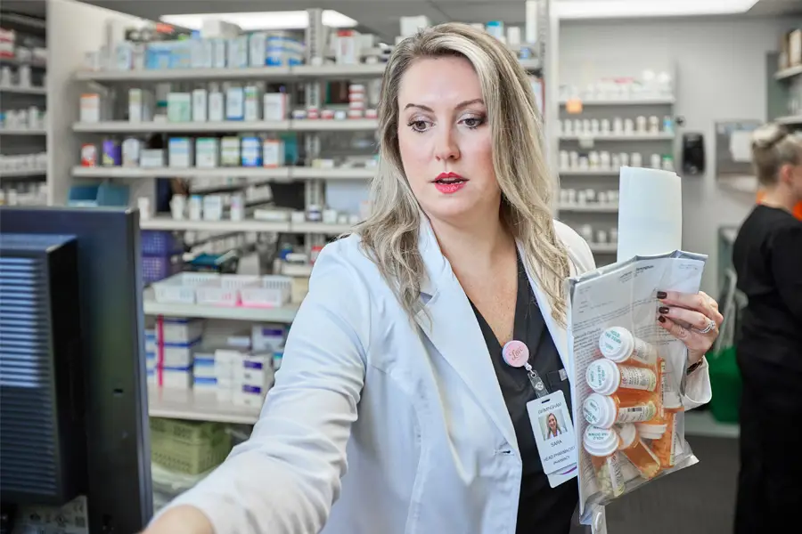 Female pharmacist in white coat holding prescription bottles and paperwork while working at computer in pharmacy