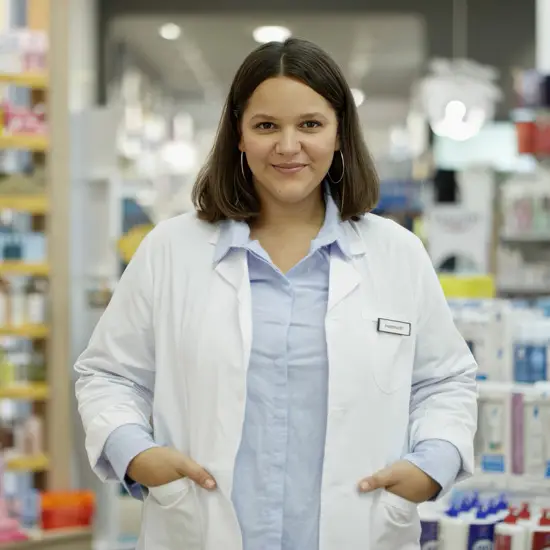 Confident female pharmacist wearing a white coat standing in a pharmacy aisle with medicines and healthcare products in the background.