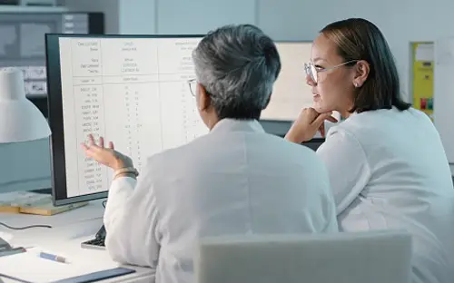 Two pharmacists in lab coats reviewing clinical data and medical charts on a large computer monitor inside a pharmacy office.