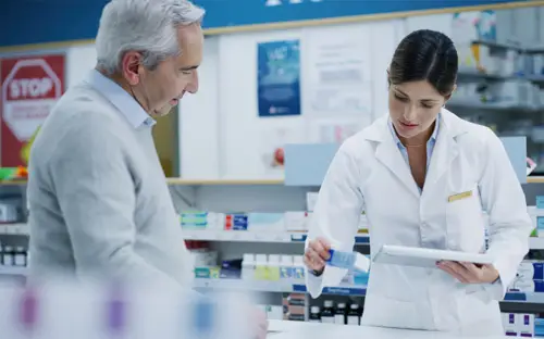 Pharmacist in white coat assisting an older male customer with prescription medication inside a pharmacy.