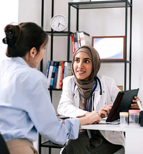 Female doctor wearing a hijab consulting with a patient in a medical office, using a digital tablet for healthcare records.
