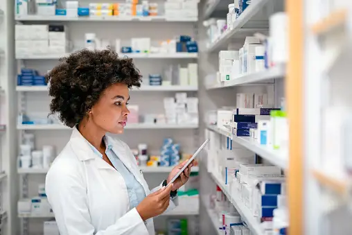 Female pharmacist in lab coat using a digital tablet while checking prescription medications on pharmacy shelves.