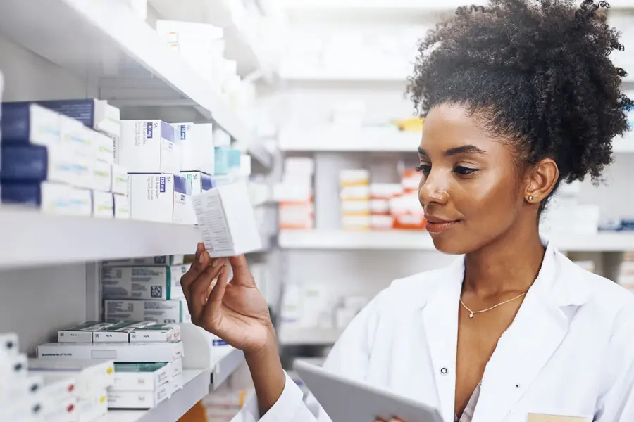 emale pharmacist in white coat checking medication box while using digital tablet in pharmacy