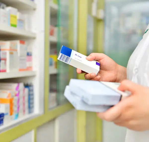 Pharmacist holding prescription medicine boxes while checking inventory on pharmacy shelves.