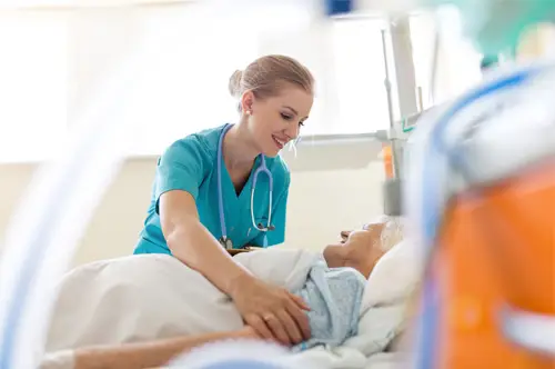 Smiling nurse in scrubs with stethoscope providing compassionate care to an elderly patient in a hospital bed.