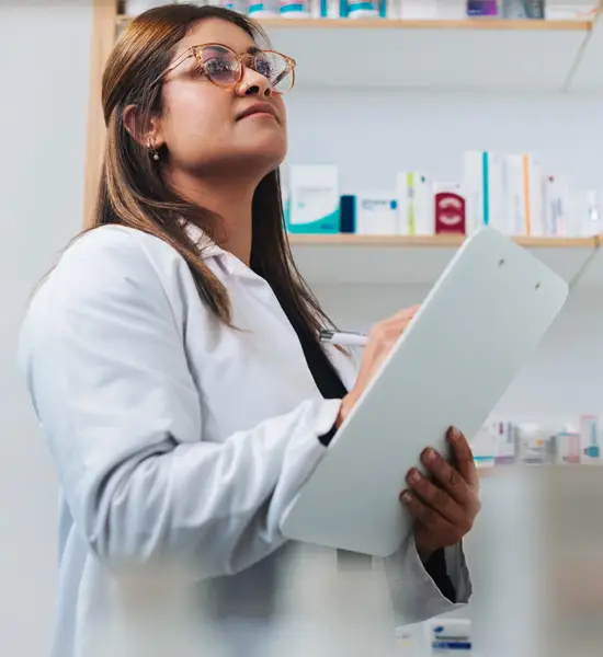 Female pharmacist in white coat holding a clipboard and taking notes while reviewing medication inventory on pharmacy shelves.
