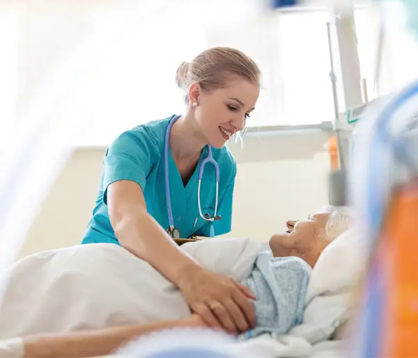 Smiling nurse in scrubs caring for elderly patient in hospital bed.