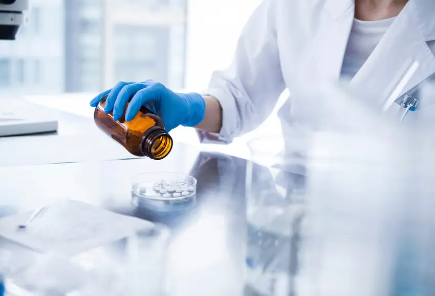 Pharmaceutical researcher in lab coat and gloves pouring pills into petri dish for testing.