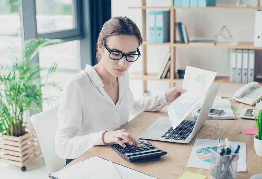 Businesswoman analyzing financial reports with calculator and laptop at office desk.