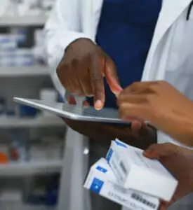 Pharmacists in lab coats reviewing medication boxes and information on a digital tablet inside a pharmacy