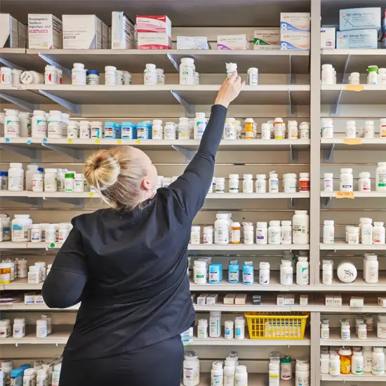 Pharmacy technician reaching for a prescription medication bottle on shelves stocked with medicine.