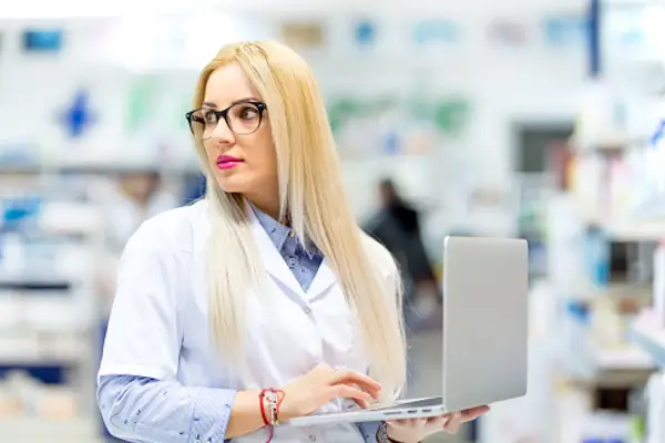 Female pharmacist with long blonde hair and glasses working on a laptop inside a bright modern pharmacy.