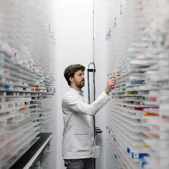 Male pharmacist in lab coat organizing prescription medications inside a modern automated pharmacy storage system.