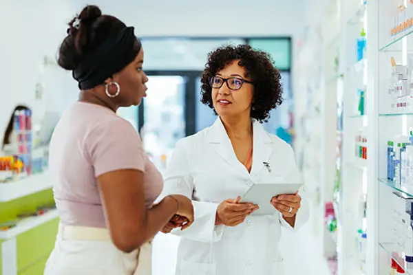 Pharmacist in white coat holding a digital tablet while consulting with a customer inside a modern pharmacy.
