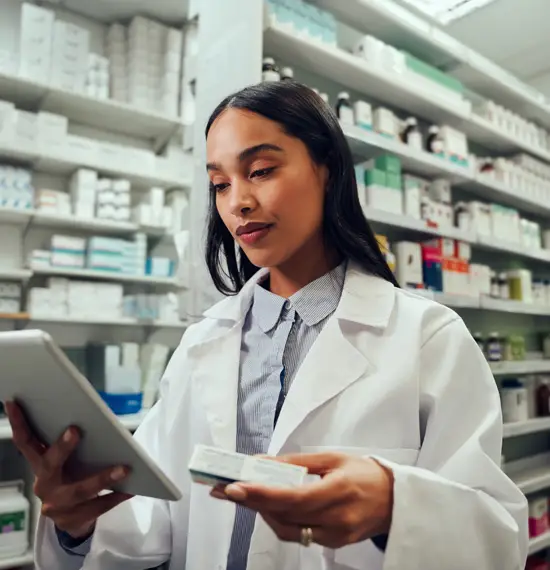 Pharmacist in white coat reviewing medication box and tablet in pharmacy aisle
