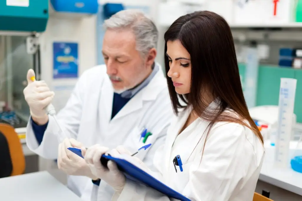 Two scientists in lab coats working together in a laboratory, one using a pipette and the other taking notes on a clipboard.