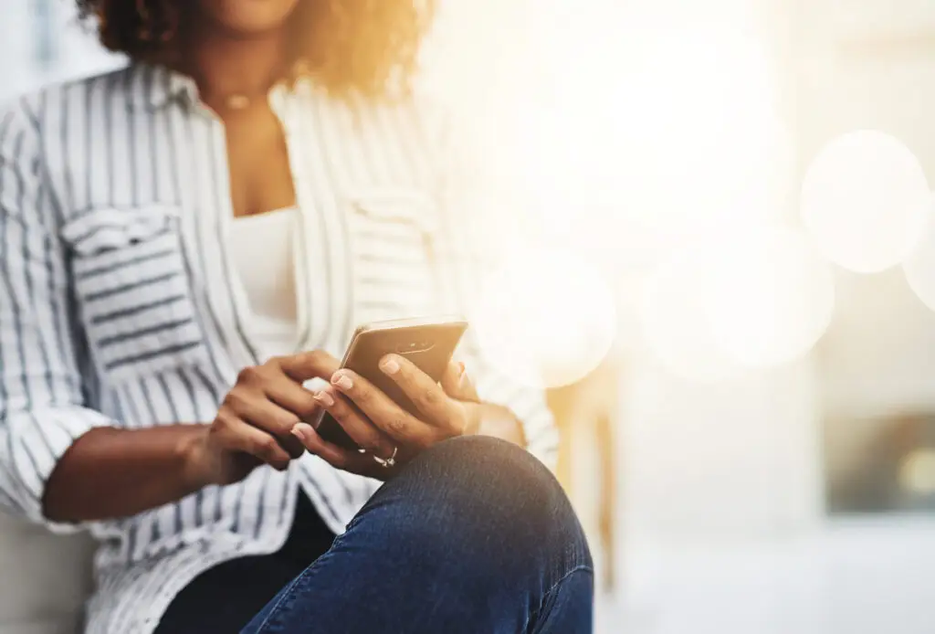 Woman in casual clothing sitting with crossed legs, using a smartphone in bright natural light.