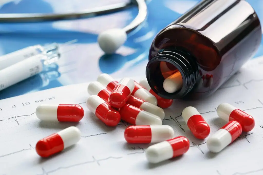 Red and white capsules spilled from a brown medicine bottle onto an ECG chart, with medical equipment in the background.