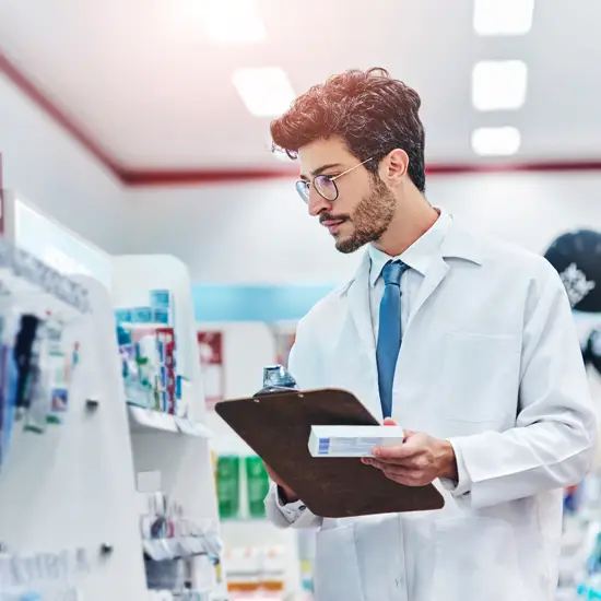 Pharmacist in a white coat holding a clipboard and a medication box while checking products on pharmacy shelves.