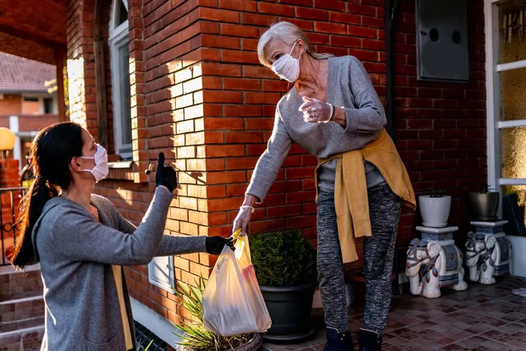 Older woman in a face mask receives a grocery bag from a younger woman wearing a mask and gloves outside a brick house.