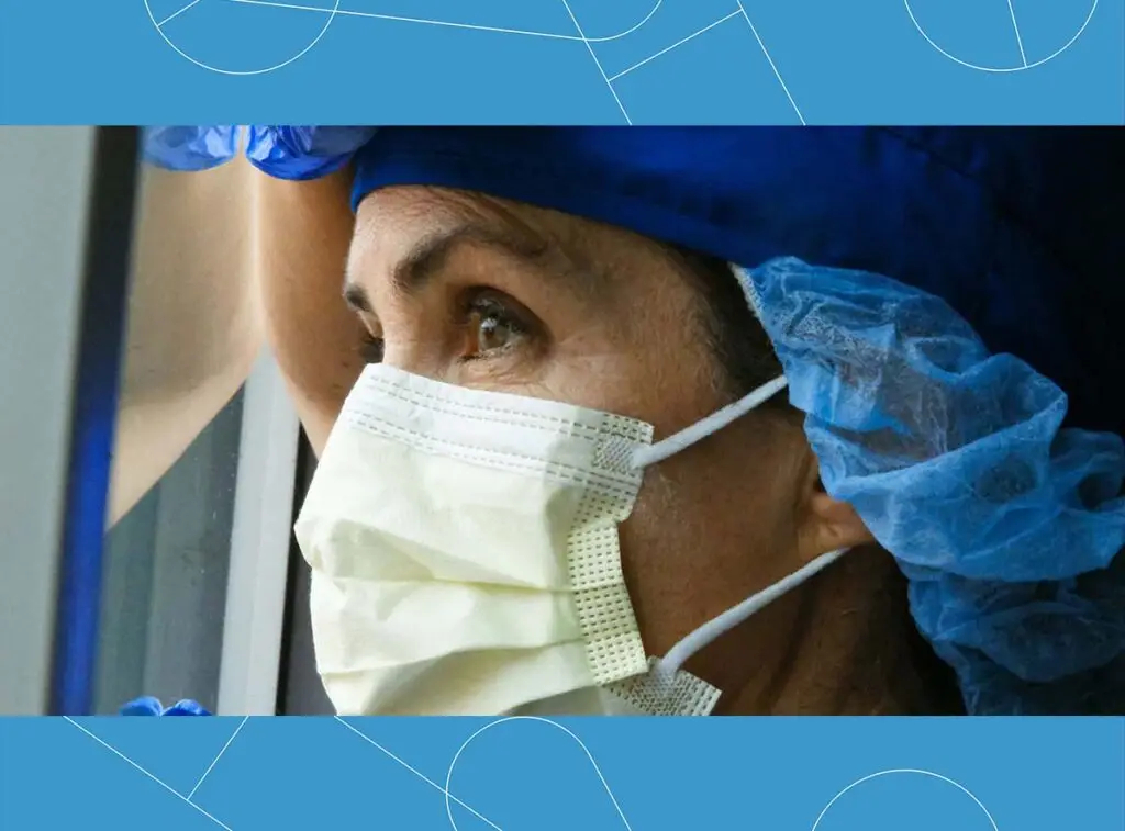 Healthcare worker wearing a surgical mask and blue hair cover looking out a window with a thoughtful expression