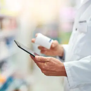 Pharmacist in a white coat holding a prescription bottle and using a digital tablet inside a pharmacy.