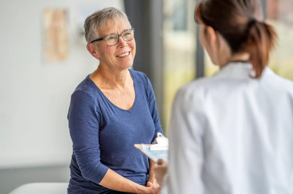 A senior woman who is dressed casually, sits up on an exam table as she attends a health check-up with her doctor.  She has a neutral expression on her face as she talks with the female doctor.  The doctor is seated across from her and has a clipboard in her hands as she takes notes throughout the appointment.